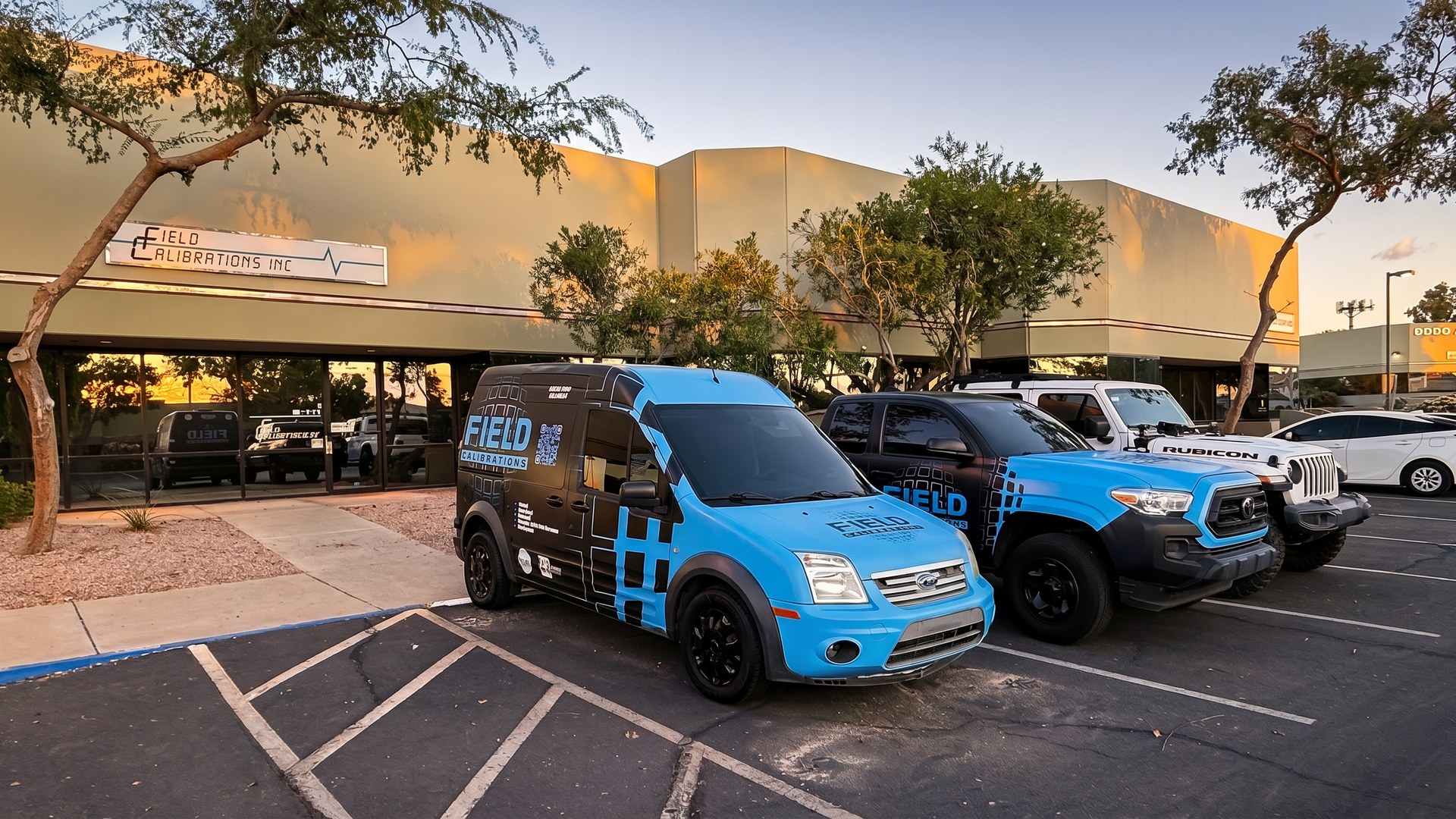 Field Calibrations fleet vehicles — wrapped Ford Transit Connect service van, Toyota Tacoma, and Jeep Rubicon — parked at the FCI Phoenix facility at golden hour, building signage visible